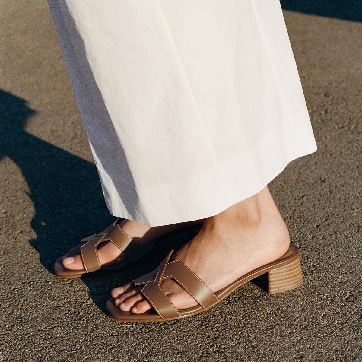 A person in white wide-leg pants and Margaux’s The MX 35 - Mocha Calf block heels stands on a sunlit pavement.