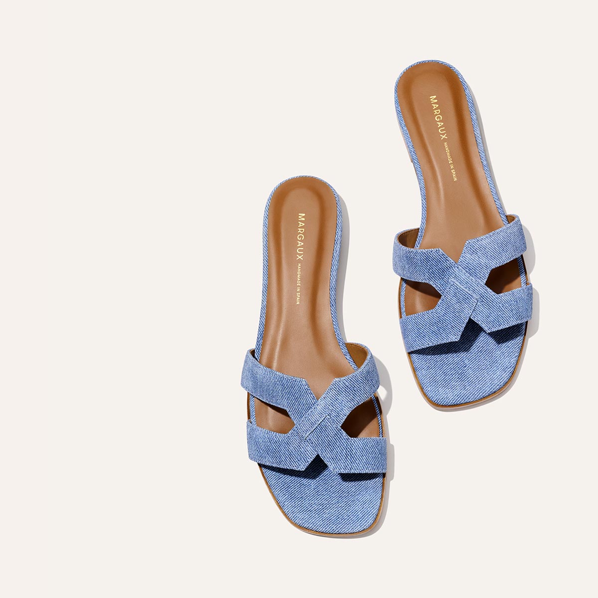 The MX Sandal - Denim Suede by Margaux features blue slip-on crisscross straps, tan insoles, a flat sole, and a square open toe, shown on a light background.