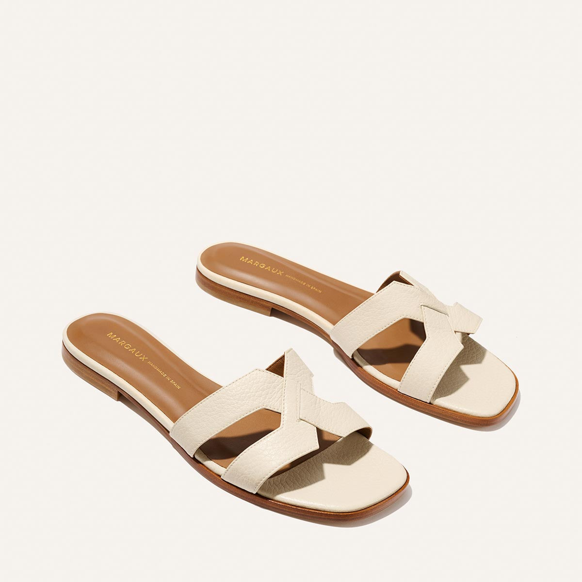 The Margaux MX Sandal in Ecru Tumbled Nappa features stylish, flat slip-ons with tan soles and cream crisscross luxe leather straps—your go-to everyday sandal, shown on a white background.