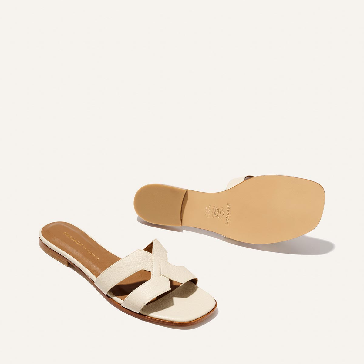 The MX Sandal - Ecru Tumbled Nappa by Margaux features ivory crisscross fabric straps, tan insoles, and a brown sole—one is shown from the side while the other highlights the smooth sole. Perfect for everyday wear.