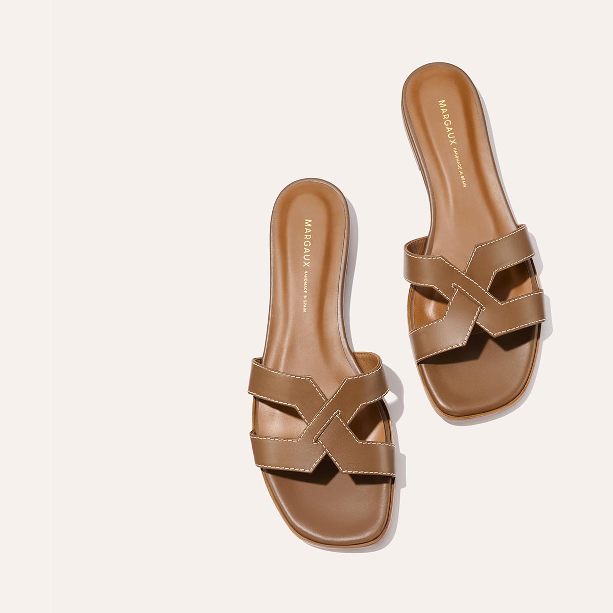 The MX Sandal - Mocha Calf by Margaux features brown slip-on cross-straps, white stitching, flat square toes, and the Margaux logo on the insole, shown against a light background.