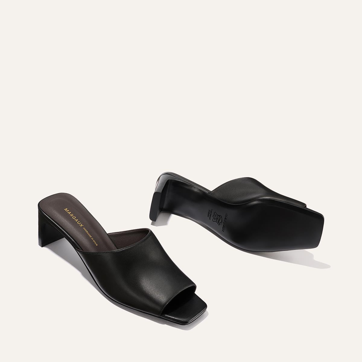 Margaux's The Naomi 50 - Black Nappa heeled mules, featuring a square toe and soft black leather upper, are displayed on a light background: one upright showing the top and side, the other on its side revealing the sole.