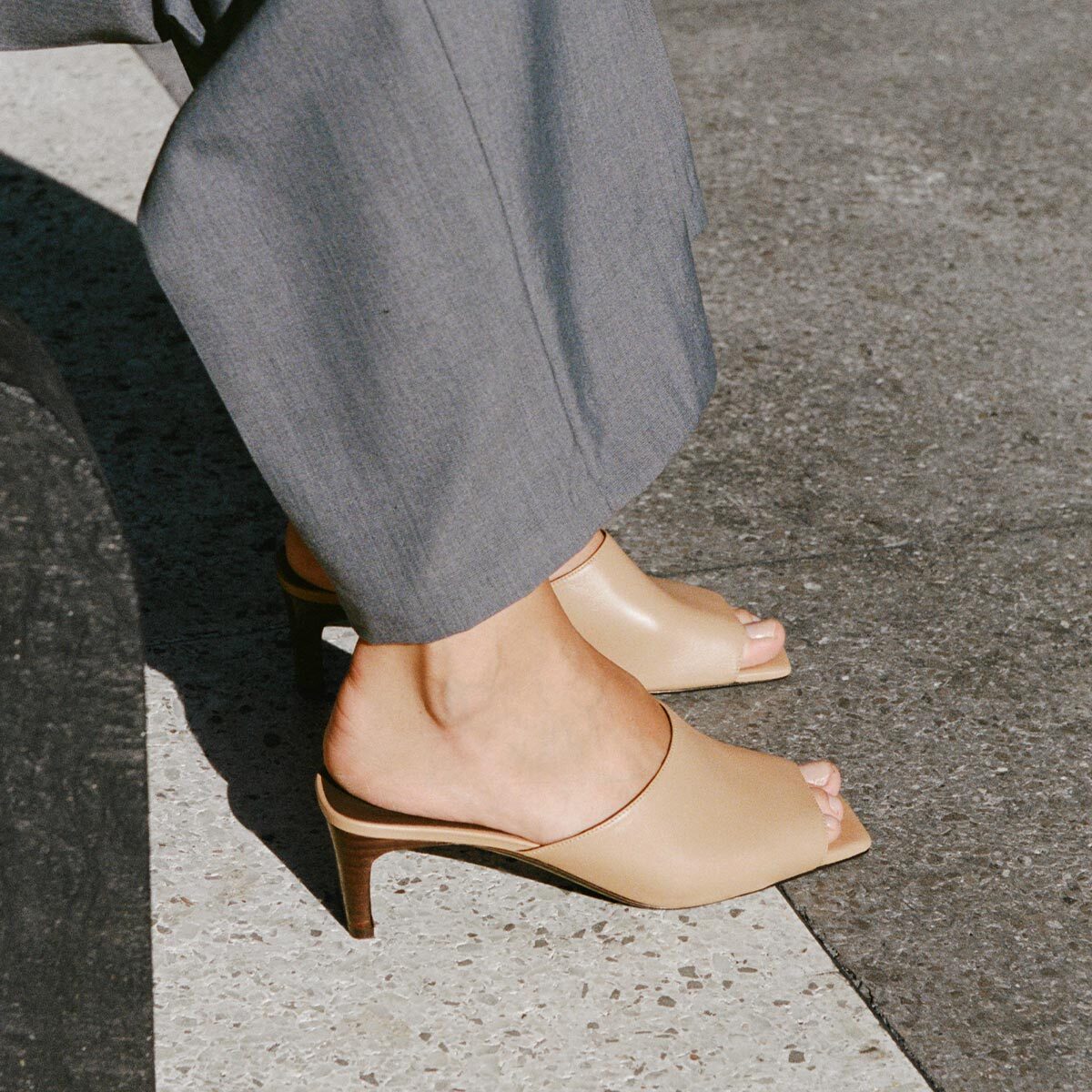 Beige open-toe high-heeled shoes worn with gray pants on a pavement.