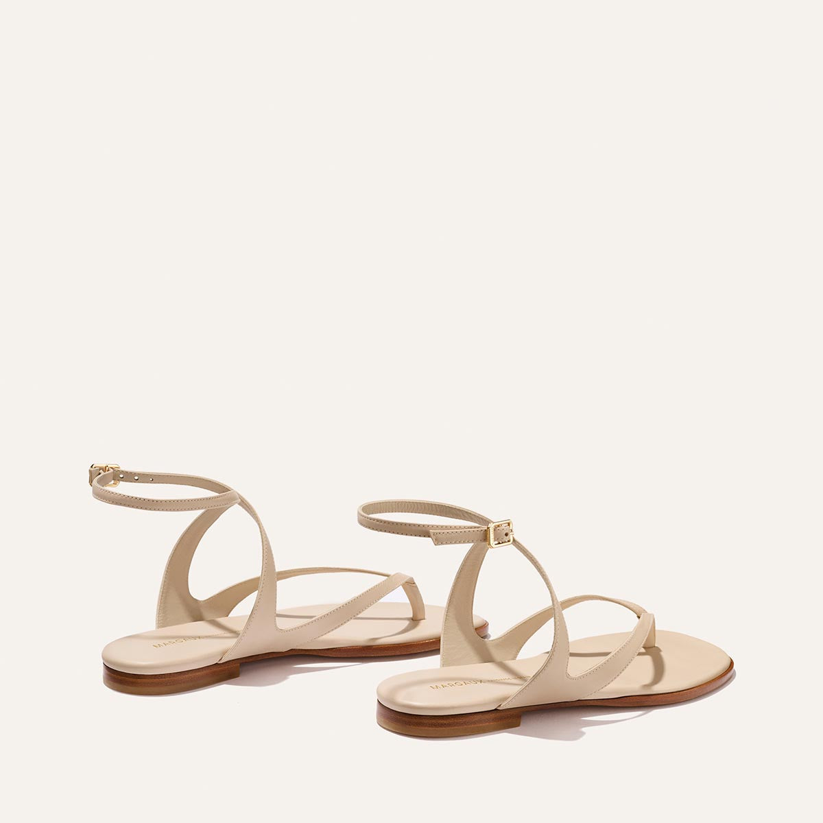 The Palermo Sandal - Magnolia Nappa by Margaux features thin, crisscrossing beige straps and ankle buckles with a cushioned insole—perfect for summer, shown on a light background from a rear side angle.