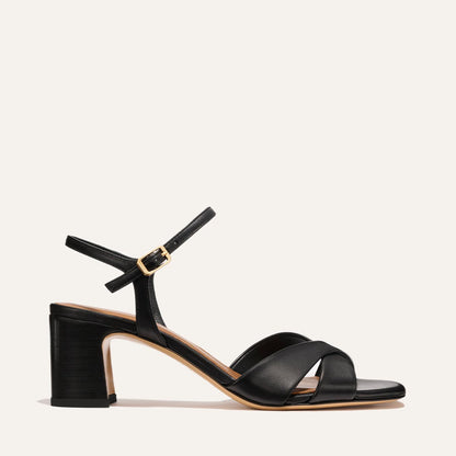 The Paloma Sandal by Margaux in Black Nappa features a mid-height block heel, crisscross toe straps, and an ankle strap with a gold buckle, shown on a plain light background.