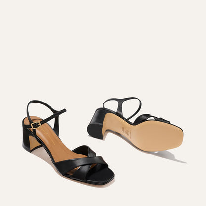 The Paloma Sandal by Margaux in Black Nappa features open toes, crisscross straps, a buckle ankle strap, low block heels, and light tan soles, shown on a light background.
