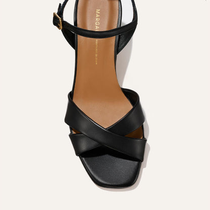 The Paloma Sandal - Black Nappa by Margaux features black crisscrossed straps, a square toe, and an ankle strap with a buckle. It has a tan insole accented by gold Margaux branding.