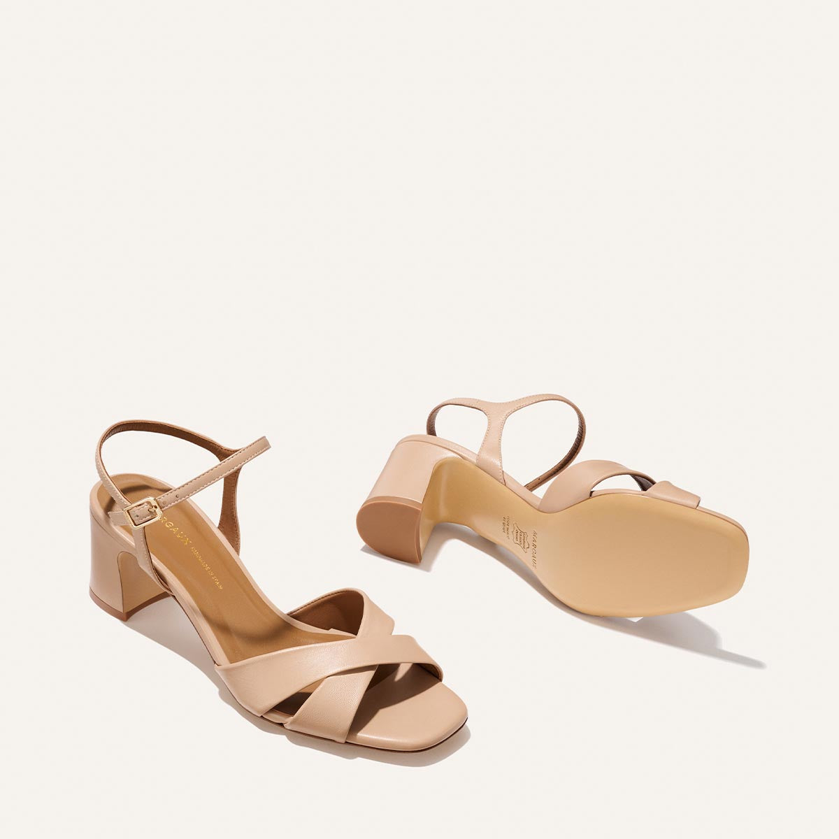 The Paloma Sandal - Dune Nappa by Margaux features beige block heels, open toes, crisscross front straps, and ankle straps with side buckles; one sandal is upright while the other rests on its side.