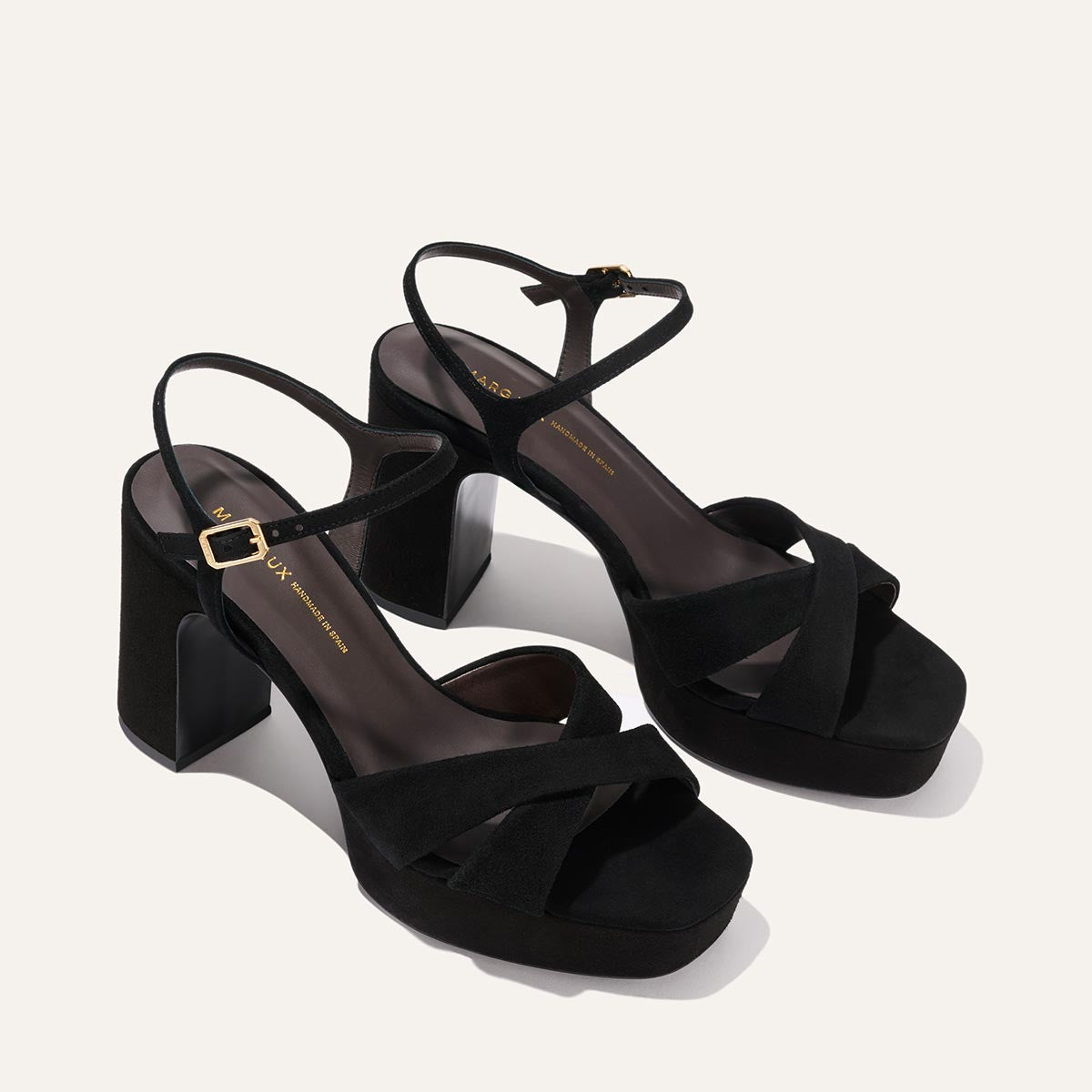 The Paloma Platform by Margaux in black suede features chunky block heels, crisscross and ankle straps with a gold buckle, plus a padded platform for comfort—perfect heeled event sandals, shown on a light background.