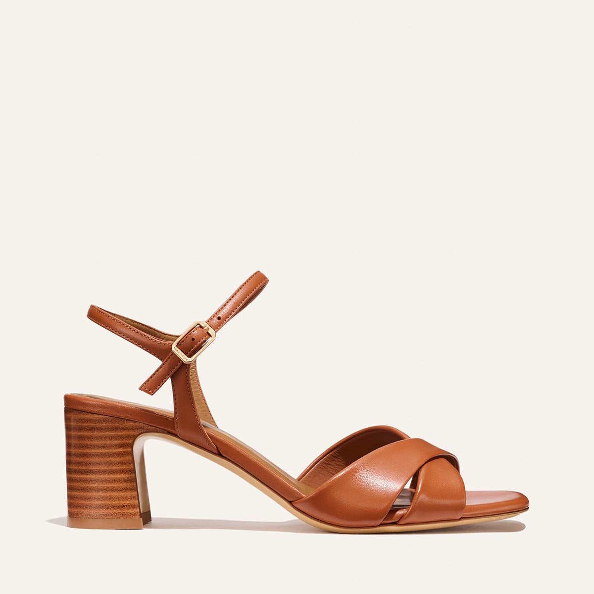 The Paloma Sandal by Margaux in Saddle Nappa features a tan leather finish, block stacked heel, open toe, crisscross front straps, and an ankle strap with a gold buckle. Displayed against a plain light background.