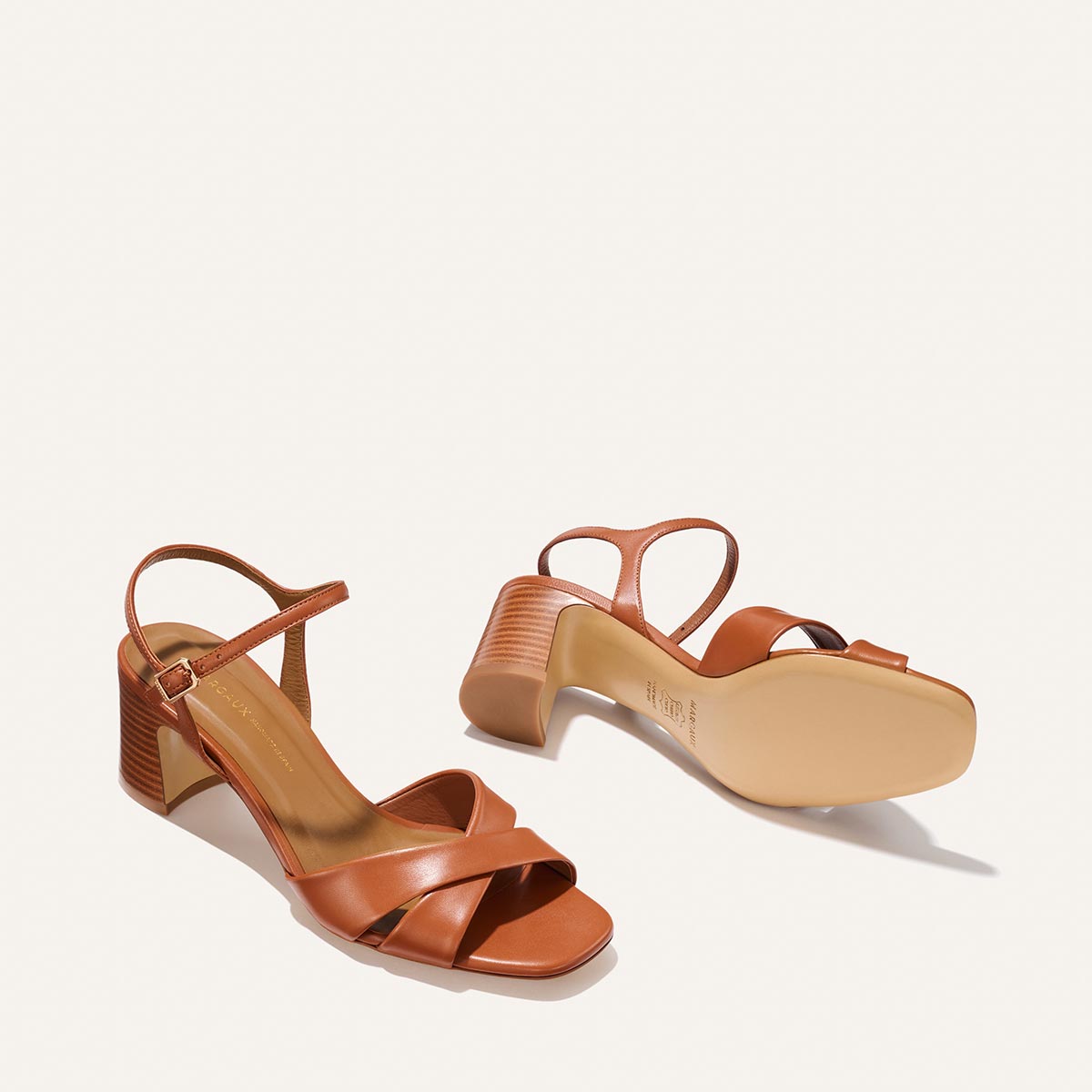 The Paloma Sandal - Saddle Nappa by Margaux features brown ankle-strap heeled sandals with crisscross front bands, block heels, and a smooth sole, shown on a light background.