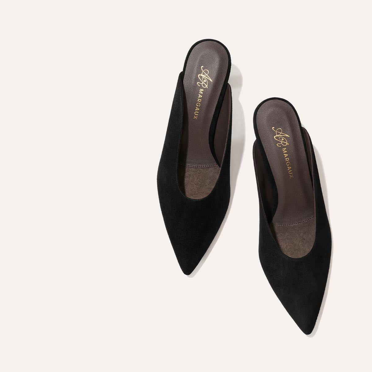 A pair of Alison Roman x Margaux: The Parlor Mule - Black Suede slip-on shoes by Margaux, featuring pointed toes and low heels, is displayed from above on a white background.