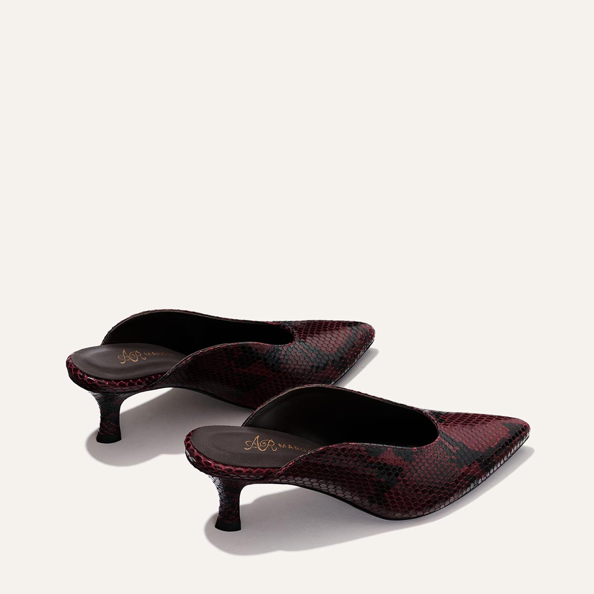 The Alison Roman x Margaux: The Parlor Mule in Burgundy Python Embossed by Margaux—a pair of heeled mules featuring a red and black snakeskin pattern, pointed toes, and low, curved heels—are positioned on a light background.