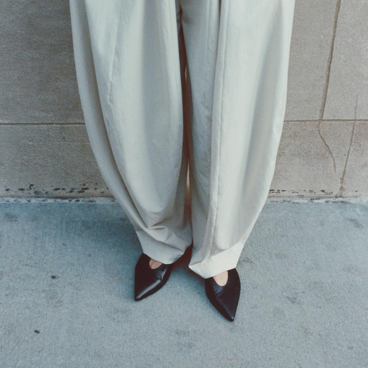 A person wearing loose, light beige pants and margaux-development’s The Parra - Black Nappa flats stands on a concrete sidewalk next to a tan stone wall.