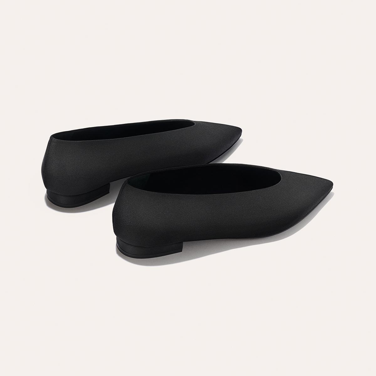 Two sleek, minimalist black satin flats—The Parra by margaux-development—are placed on a light background, one slightly in front of the other, highlighting their pointed-toe design.