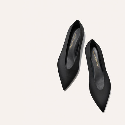 A pair of The Parra - Black Satin flats by margaux-development, featuring pointed toes and branded insoles, displayed on a light off-white background.