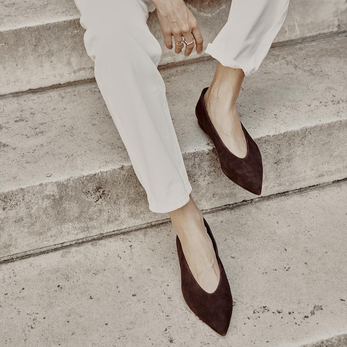 A person wearing white pants and The Parra - Espresso Suede flats by margaux-development sits on concrete steps, one hand resting on their knee and multiple rings adorning their fingers.