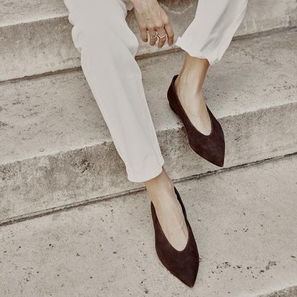 A person wearing white pants and The Parra - Espresso Suede flats by margaux-development sits on concrete steps, one hand resting on their knee and multiple rings adorning their fingers.