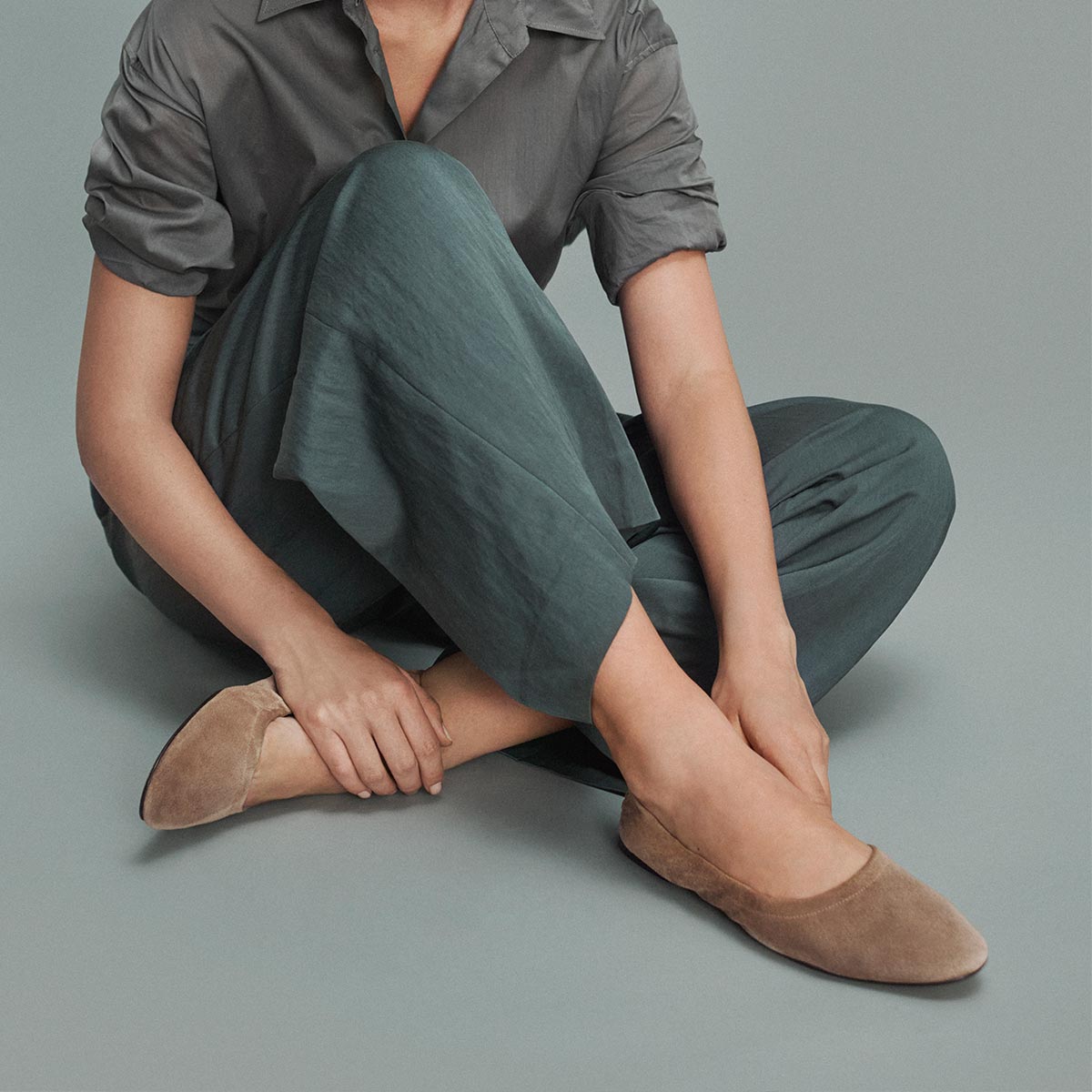 A person sits on the floor in dark teal pants and a short-sleeve shirt, adjusting Margaux's The Pascale ballet flat in Cappuccino Suede, which features an elasticated back, against a plain grey background.