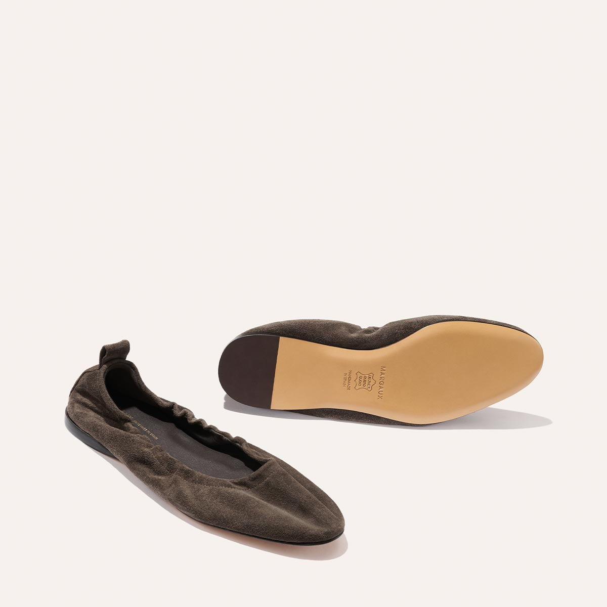 A pair of The Pascale ballet flats in English Brown Suede by Margaux, crafted from supple leather with an elasticated back—one shoe resting on its side, the other displaying the tan sole adorned with Margaux branding.