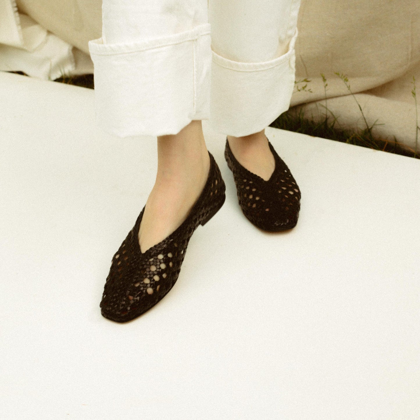 Person wearing white pants and black woven flats standing on a light-colored surface with some grass visible in the background.