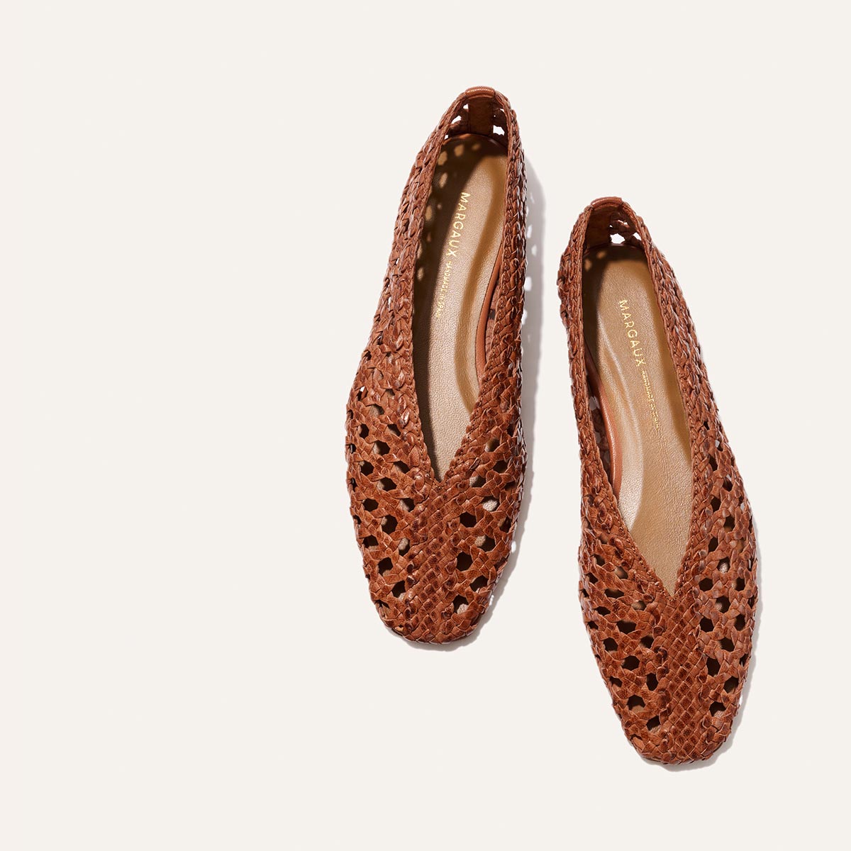 The Margaux “The Woven Paz - Saddle Leather” flats feature brown handwoven leather with pointed toes, a cut-out pattern, and tan insoles, shown on a plain white background.