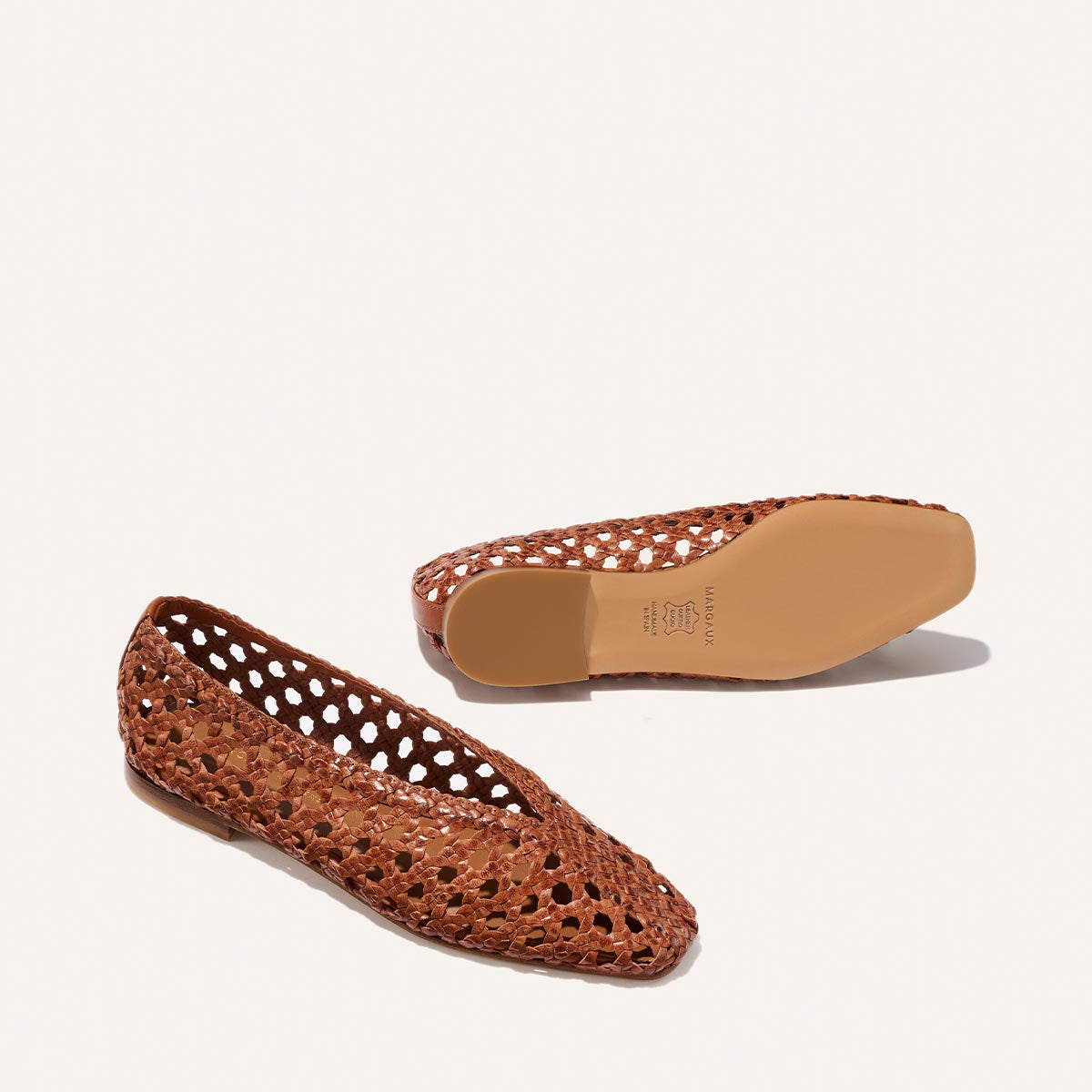 The Woven Paz by Margaux in saddle leather features luxuriously soft, handwoven brown flats with a pointed toe and cutout design. One shoe is shown from the side, the other reveals the tan sole, both set against a plain white background.