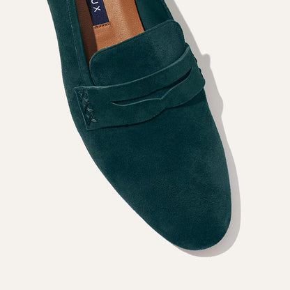 A close-up of The Penny - Emerald Suede by Margaux, a dark green Italian suede loafer with a rounded toe, penny strap detail, tan inner lining, and unlined construction for a glove-like fit, shown against a light background.