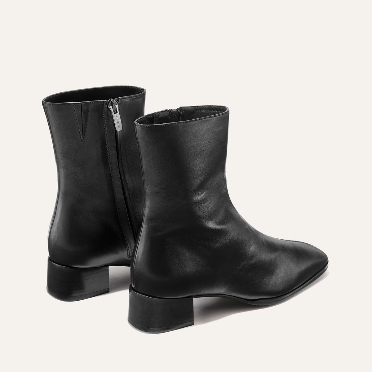The Phoebe Boot - Black Calf by Margaux: a pair of black leather ankle boots featuring low block heels, side zippers, and a smooth, slightly shiny finish. Crafted using the artisanal sacchetto technique for exceptional flexibility and comfort, these boots are displayed on a plain light background.