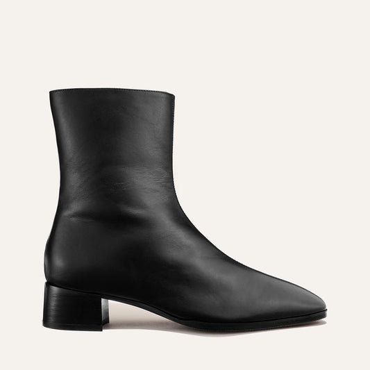 Introducing The Phoebe Boot - Black Calf by Margaux: a black leather ankle boot featuring a low block heel and gently pointed toe, expertly crafted with the artisanal sacchetto technique for exceptional flexibility and comfort. Displayed in profile against a plain off-white background.