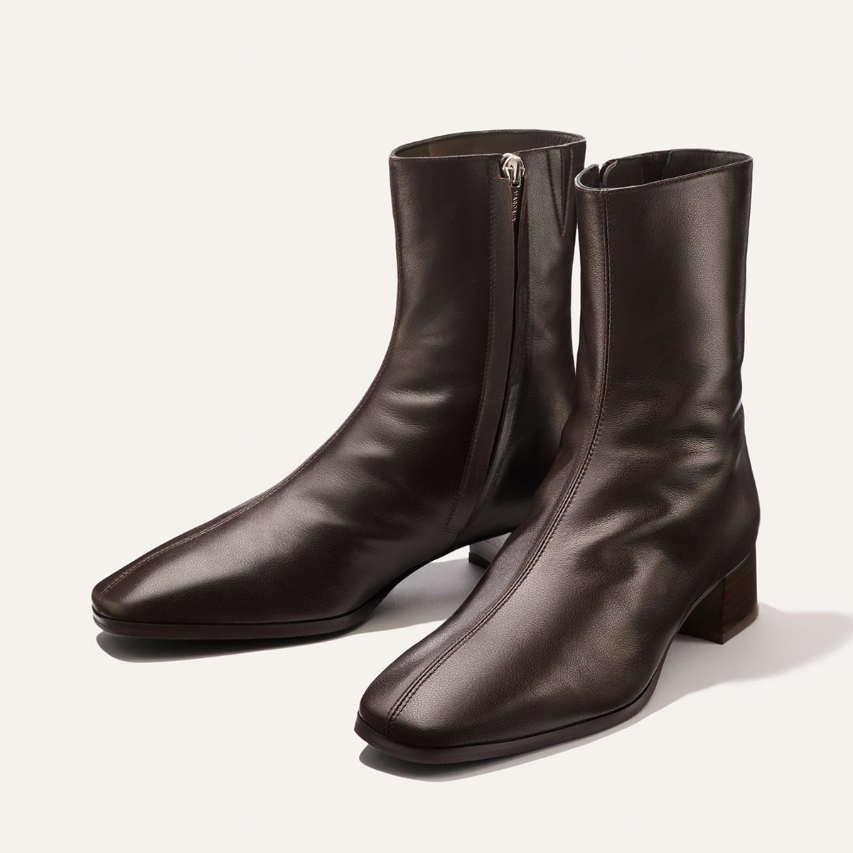 The Phoebe Boot in Dark Chocolate Calf by Margaux—a pair of dark brown leather ankle boots featuring side zippers, low block heels, and a slightly square toe—are expertly crafted using the artisanal sacchetto technique for enhanced flexibility and comfort, shown here on a light background.