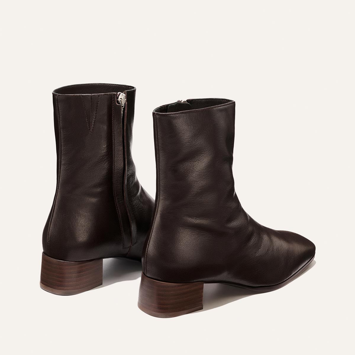 The Phoebe Boot - Dark Chocolate Calf by Margaux is a pair of rich dark chocolate leather ankle boots with side zippers and low brown wooden heels, expertly crafted using the artisanal sacchetto technique for superior flexibility and comfort. Featuring a padded insole, these boots are showcased on a plain white background.