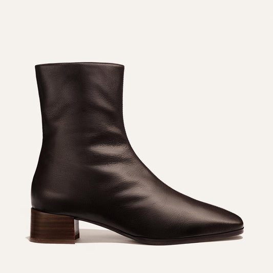 A single dark brown leather ankle boot with a pointed toe and a stacked wood-look block heel, shown in profile against a light cream background.