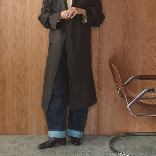 A person wearing a long dark coat, cuffed jeans, and The Phoebe Boot in Dark Chocolate Calf by Margaux stands beside a brown chair against a wooden wall. Their face is not visible.