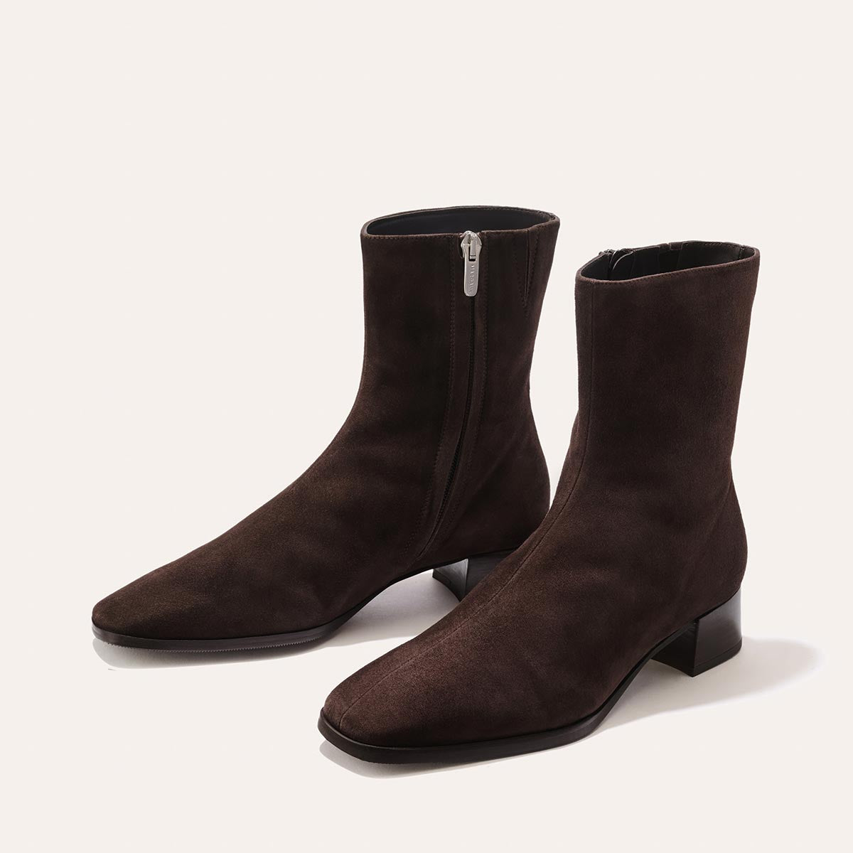 The Phoebe Boot - Molasses Suede by Margaux is a pair of dark brown suede ankle boots with side zippers and low block heels, expertly crafted using the artisanal sacchetto technique for exceptional flexibility and finished with a padded insole, shown on a light background.