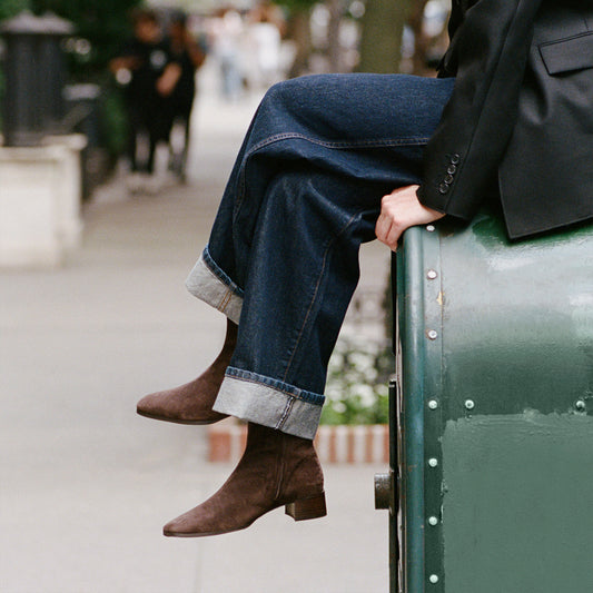 A person wearing cuffed jeans and The Phoebe Boot in Molasses Suede by Margaux, crafted with the artisanal sacchetto technique, sits on a green metal structure outdoors with one leg crossed over the other. The background is blurred, revealing a city sidewalk.