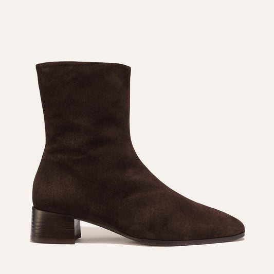 Introducing The Phoebe Boot in Molasses Suede by Margaux—a dark brown suede ankle boot with a low, block heel and almond-shaped toe. Crafted using the artisanal sacchetto technique for outstanding flexibility and comfort, this style features minimal stitching, a padded insole, and is showcased on a plain light background.
