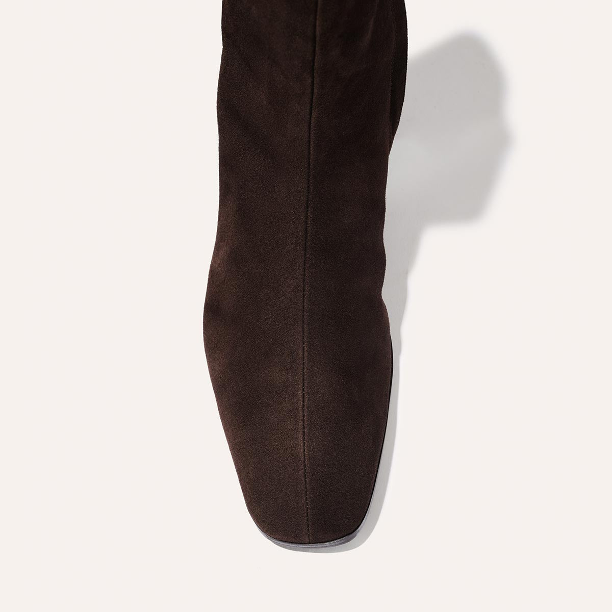 A top-down view of The Phoebe Boot - Molasses Suede by Margaux, showcasing its dark brown suede and squared toe, meticulously crafted with the artisanal sacchetto technique and finished with a padded insole, casting a soft shadow on a light background.