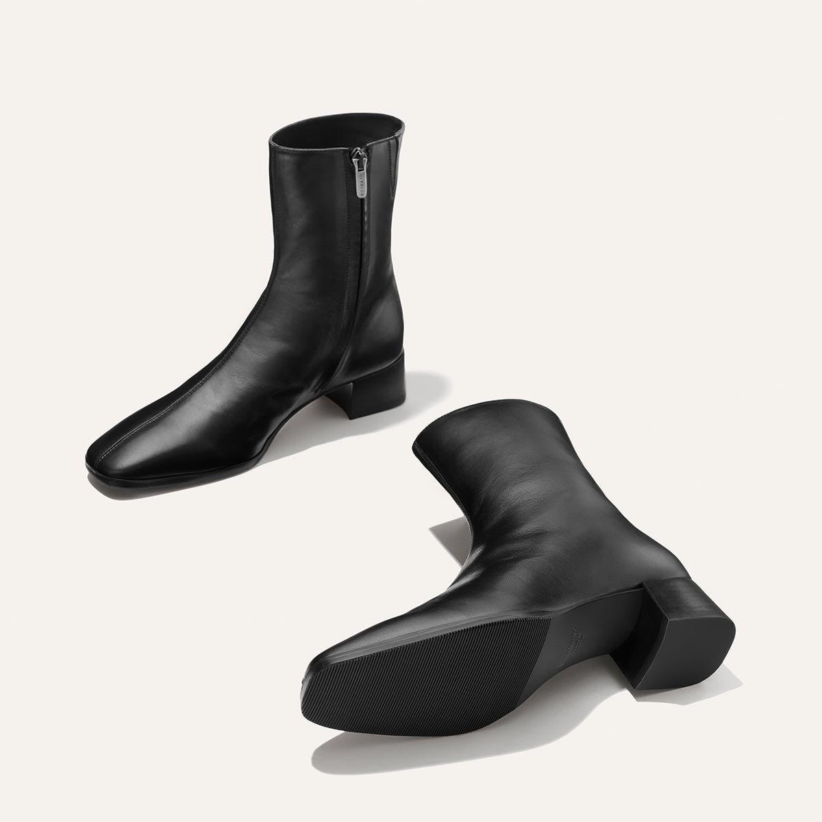 Presenting The Phoebe Boot - Black Calf by Margaux: a pair of black calf leather ankle boots, expertly crafted using the artisanal sacchetto technique. These boots feature convenient side zippers and comfortable low block heels. Shown with one boot standing upright and the other lying on its side against a white background.
