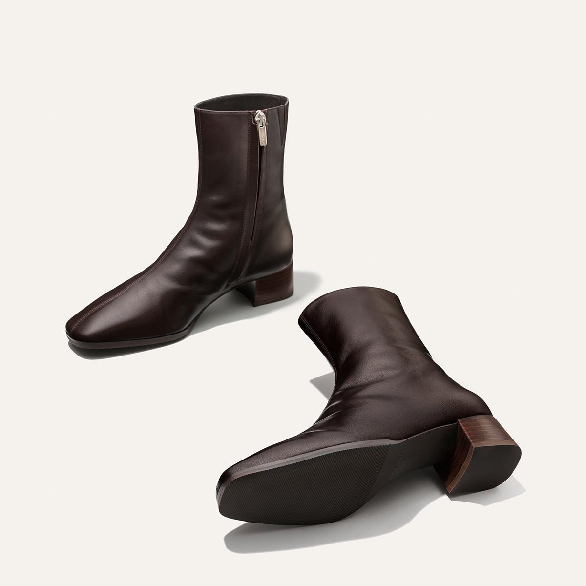 A pair of Margaux’s The Phoebe Boot in Dark Chocolate Calf—featuring dark brown leather, side zippers, and low block heels, meticulously crafted using the artisanal sacchetto technique for superior comfort—with one boot standing upright and the other resting on its side, showcased against a simple off-white background.