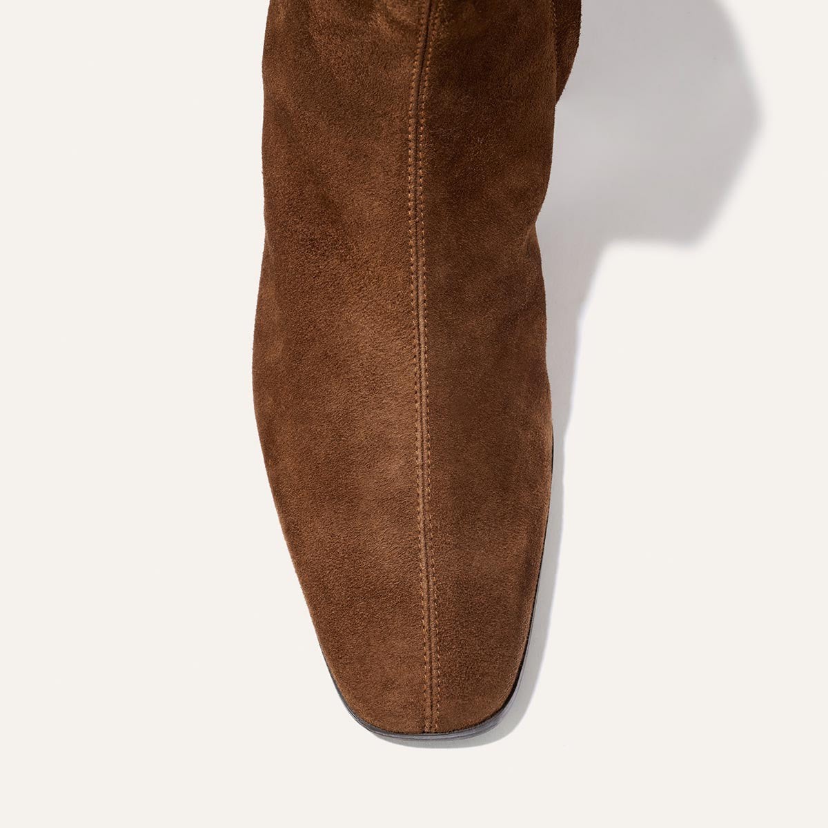 Here’s the rewritten sentence incorporating the provided product data: A top view of The Phoebe Boot - Ganache Suede by Margaux, showcasing its brown suede upper with a slightly rounded square toe, intricate stitching, and rich texture against a plain white background. Expertly crafted with the artisanal sacchetto technique, this comfortable flat boot features a padded insole for all-day comfort.