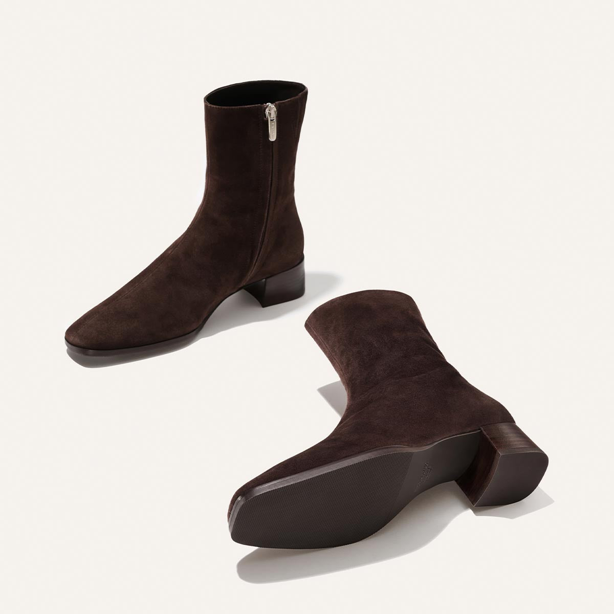 The Phoebe Boot in Molasses Suede by Margaux—a pair of dark brown suede ankle boots featuring low block heels, inner side zippers, and slightly pointed toes—are expertly crafted using the artisanal sacchetto technique for exceptional comfort and are displayed on a light background.