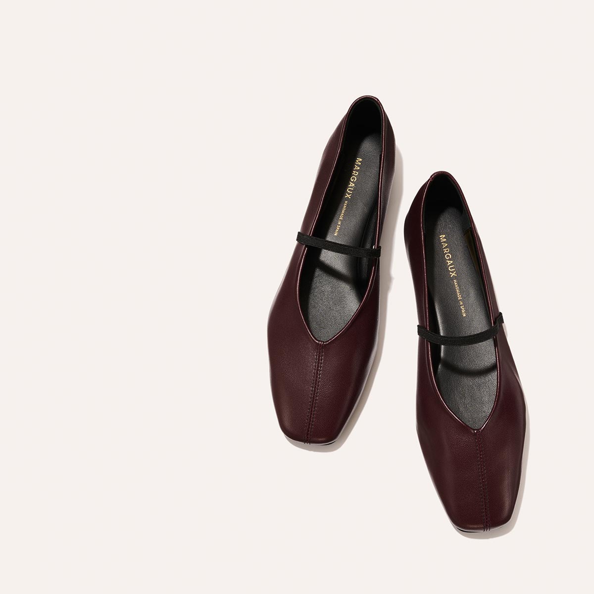 The Phoebe - Burgundy Nappa by margaux-development: burgundy leather flats with black straps and square toes, crafted with sacchetto construction, displayed on a light beige background.