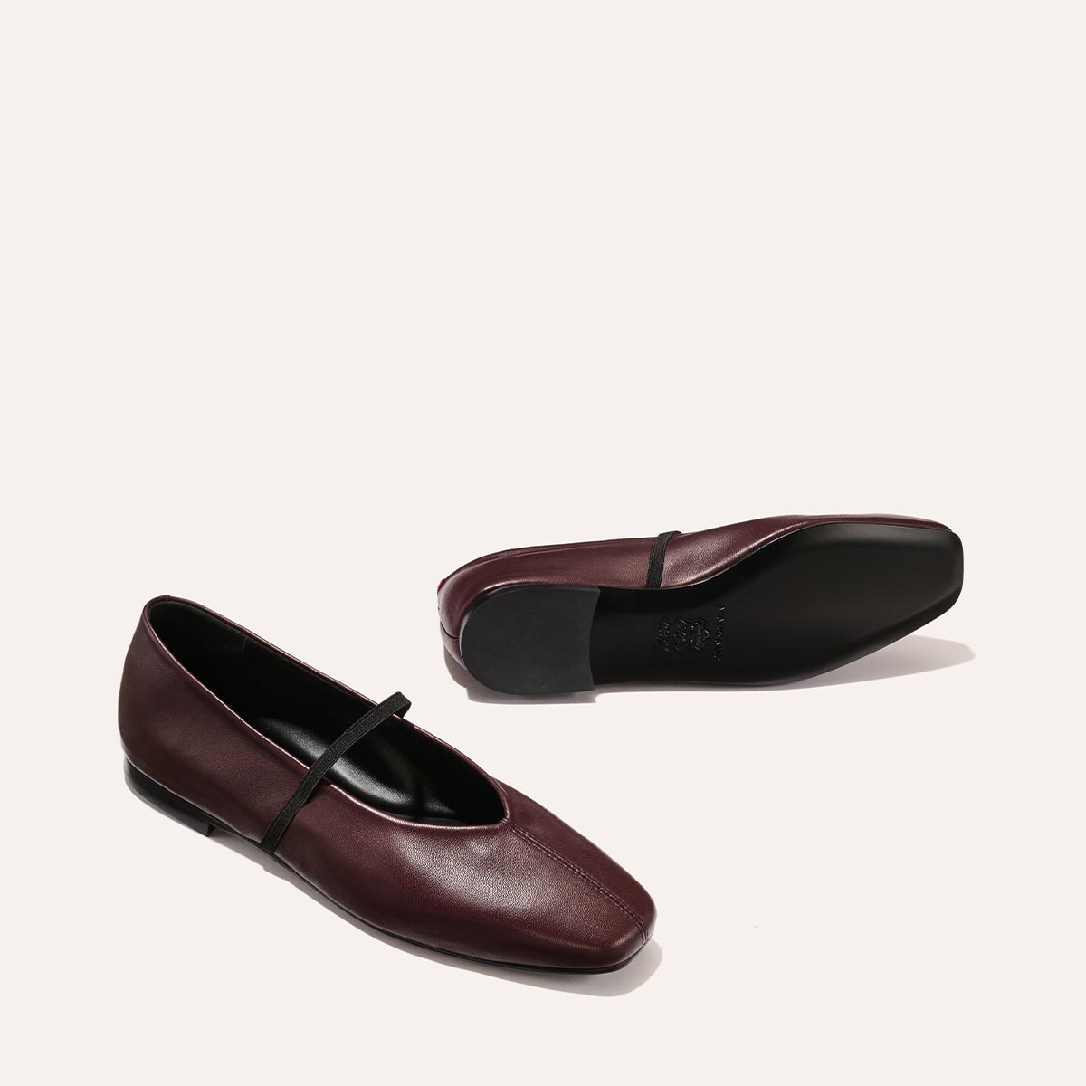 Two The Phoebe - Burgundy Nappa leather flats by margaux-development, one upright and one on its side, with black soles and elastic top straps. Sacchetto construction ensures softness. Displayed on a plain light beige background.