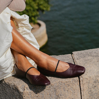 A person in a white skirt and The Phoebe - Burgundy Nappa flats by margaux-development, featuring a thin black strap, sits on a stone ledge by the water with green foliage in the background.
