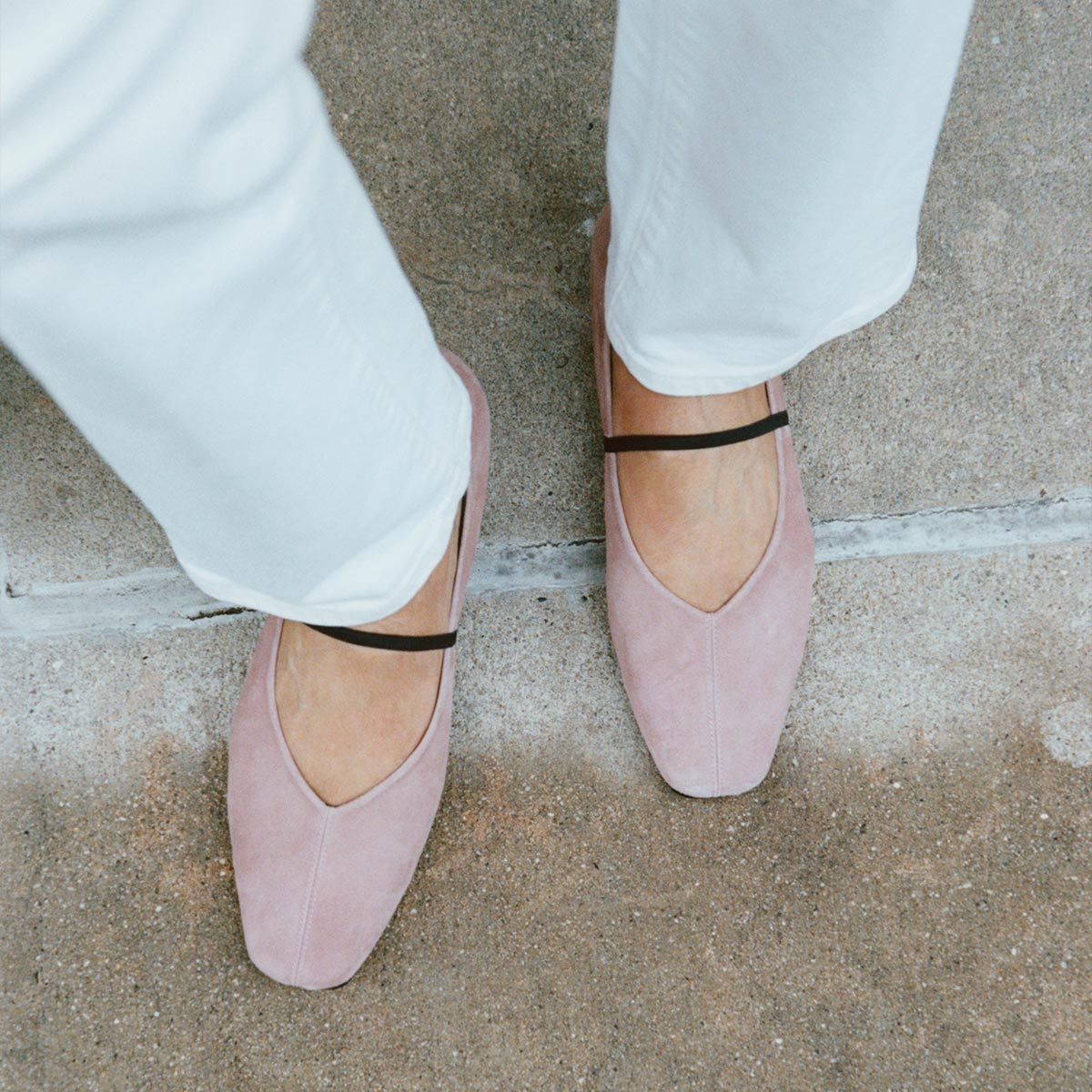 A person stands on a concrete surface wearing white pants and The Phoebe - Dusty Rose Suede flats by margaux-development, featuring square toes and black straps.