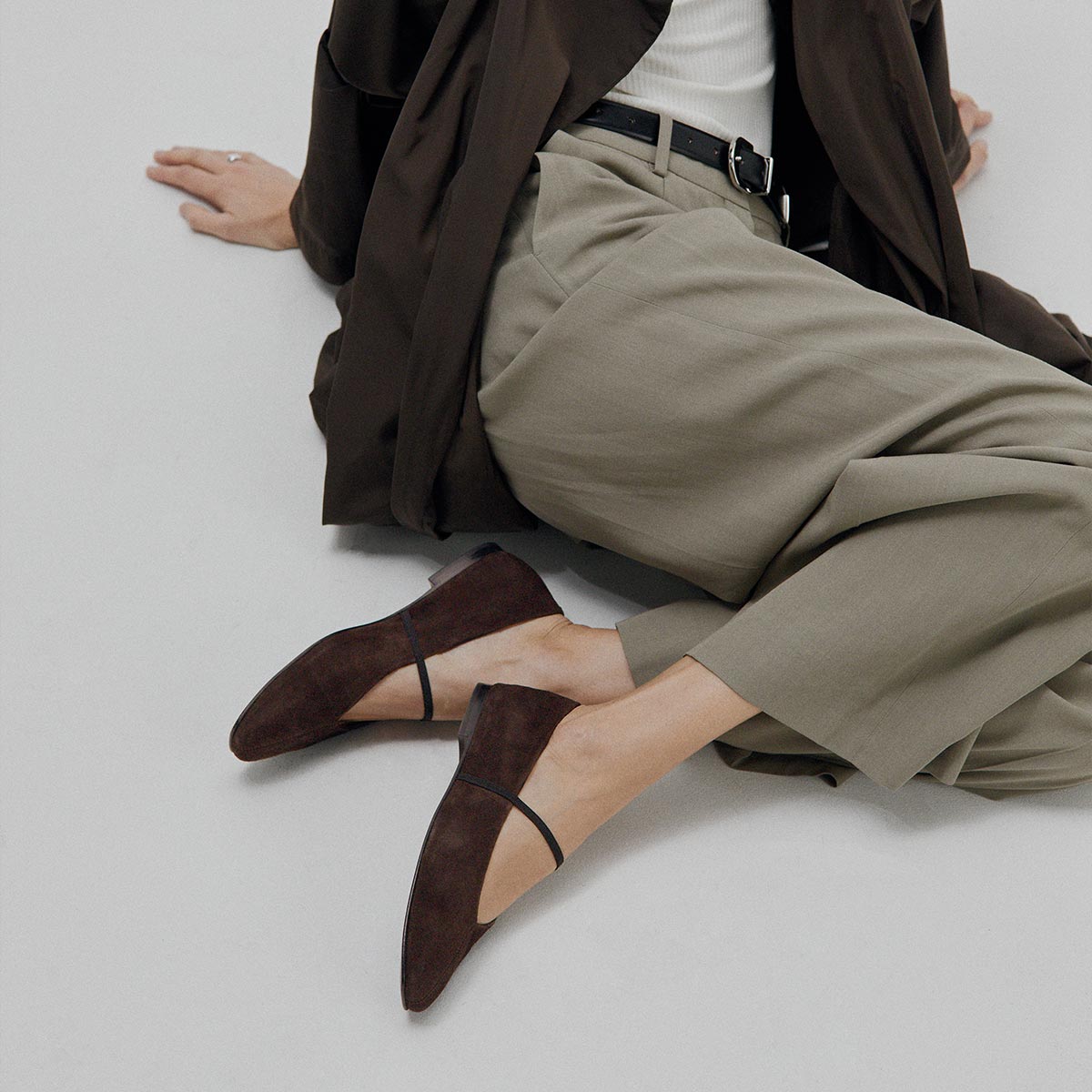 A person sits on the floor in light olive-green pants, a white top, a dark brown coat, and The Phoebe - Espresso Suede flats by margaux-development. The minimal, neutral setting enhances the effortless style.