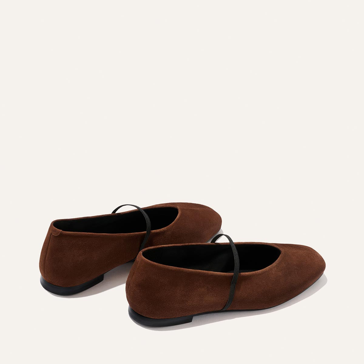 A pair of Margaux's The Phoebe - Ganache Suede ballet flats, featuring rich brown suede, black soles, and a delicate black strap across each shoe. Expertly crafted in sacchetto construction for exceptional softness, they are elegantly displayed against a plain light background.