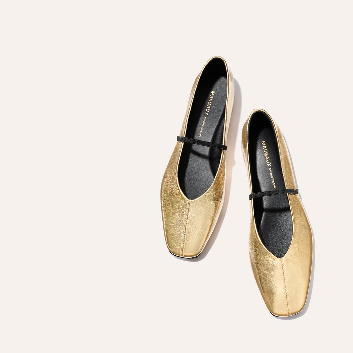The Phoebe - Gold Crinkle Nappa flats by margaux-development, featuring black straps and insoles with sacchetto construction, are placed diagonally on a light beige background.