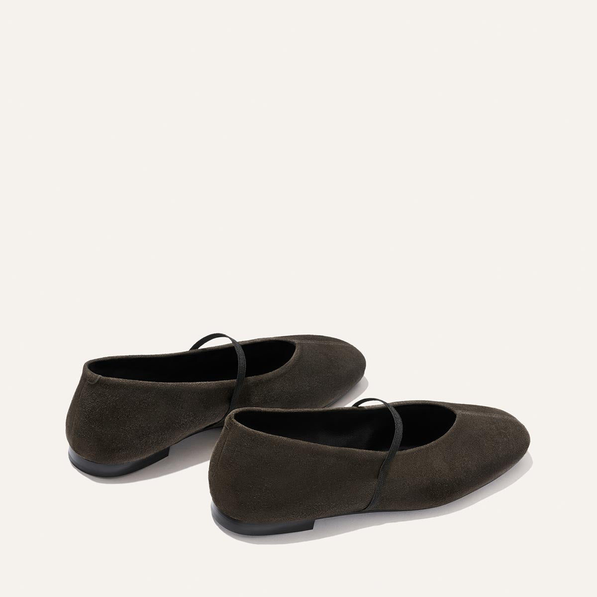 A pair of Margaux’s The Phoebe flats in graphite suede—with thin straps, black soles, and a minimalist design—expertly crafted using sacchetto construction for one of the softest flat experiences, placed on a plain light background.